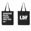 Two black canvas tote bags shown side by side on a white background. The left bag has white text reading 'ADVANCING RACIAL JUSTICE SINCE 1940' with a small LDF logo below.