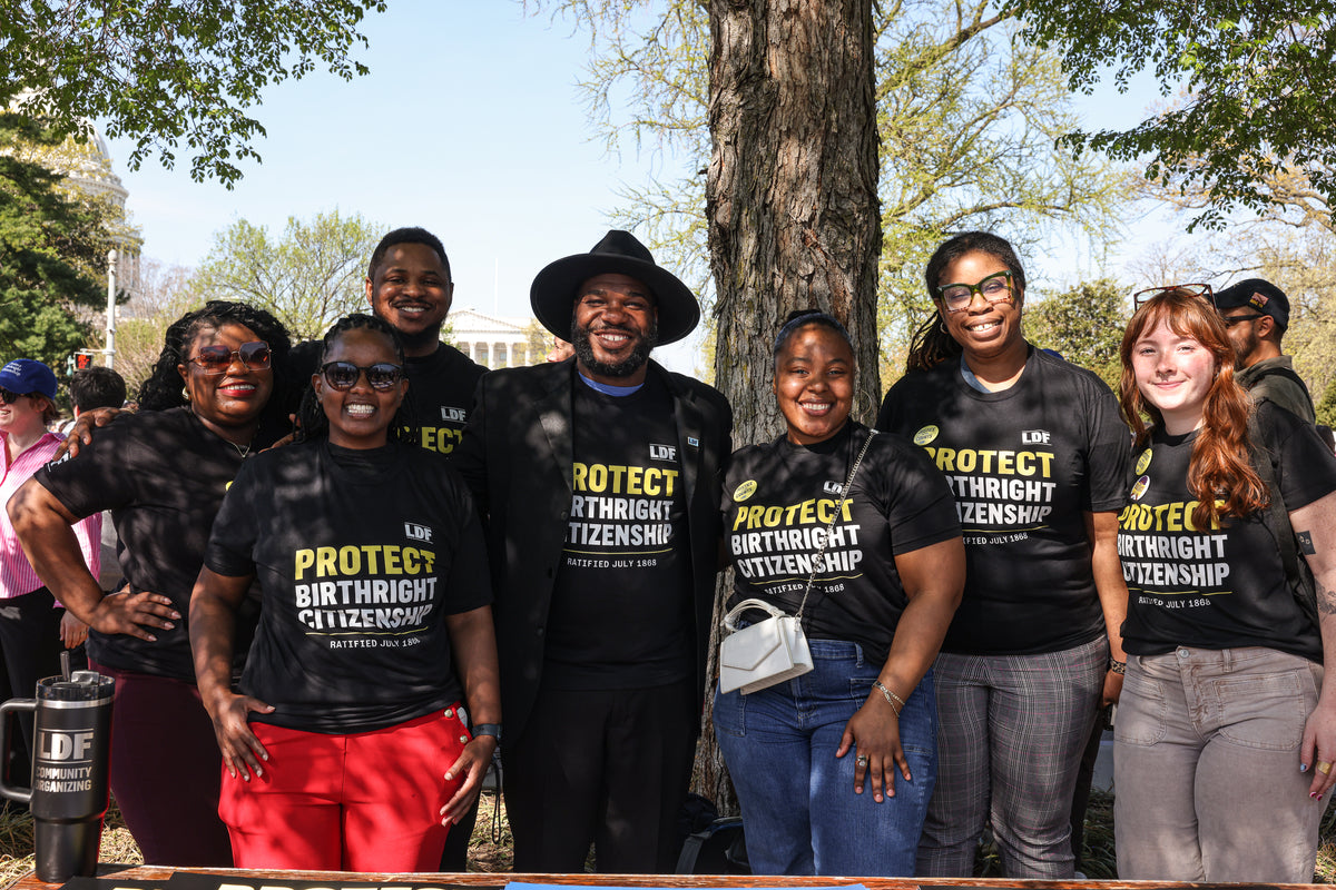 A group of seven people standing outdoors in front of a tree, wearing matching black t-shirts with yellow and white text that reads 'PROTECT BIRTHRIGHT CITIZENSHIP' and 'LDRB'.