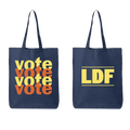 Two navy blue canvas tote bags shown side by side on a white background. The left bag features the word 'vote' printed repeatedly in a stacked, bold yellow and orange font.